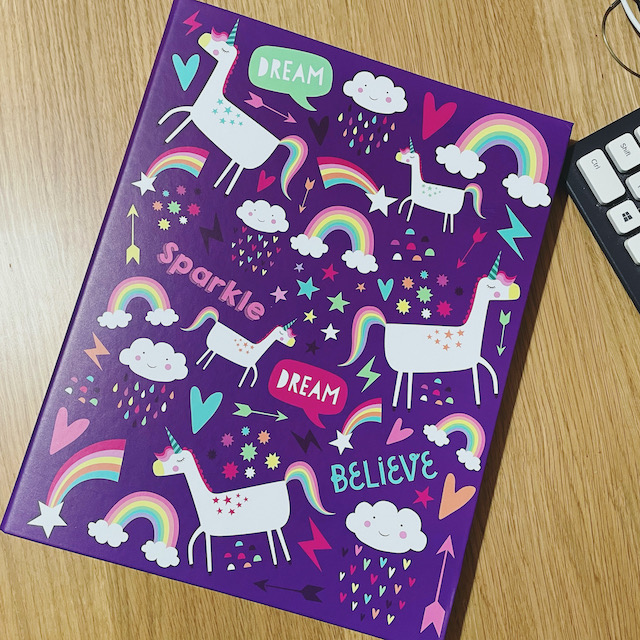 A purple ring-binder covered in rainbows, unicorns, and happy little clouds, with wholesome messages to dream, believe, and sparkle