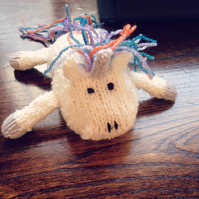 A tiny hand-knitted unicorn with mane and tail made from rainbow sock yarn, splatted on a pub table