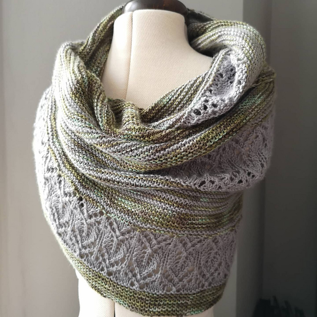 A hand-knitted shawl/scarf in muted green with grey lace panels, wrapped around a tailor's dummy.