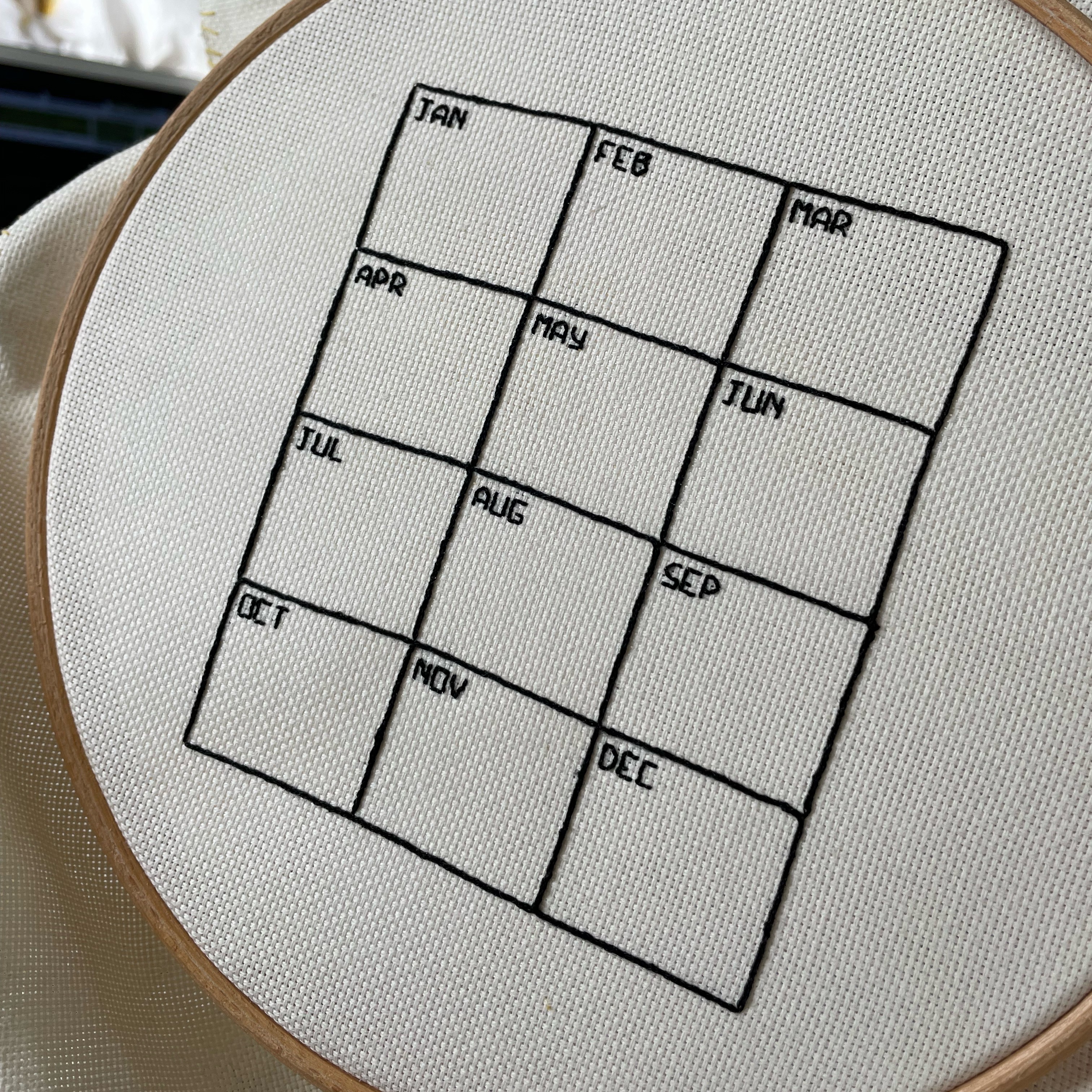 A calendar stitched onto cream aida embroidery cloth, three months across by four months down